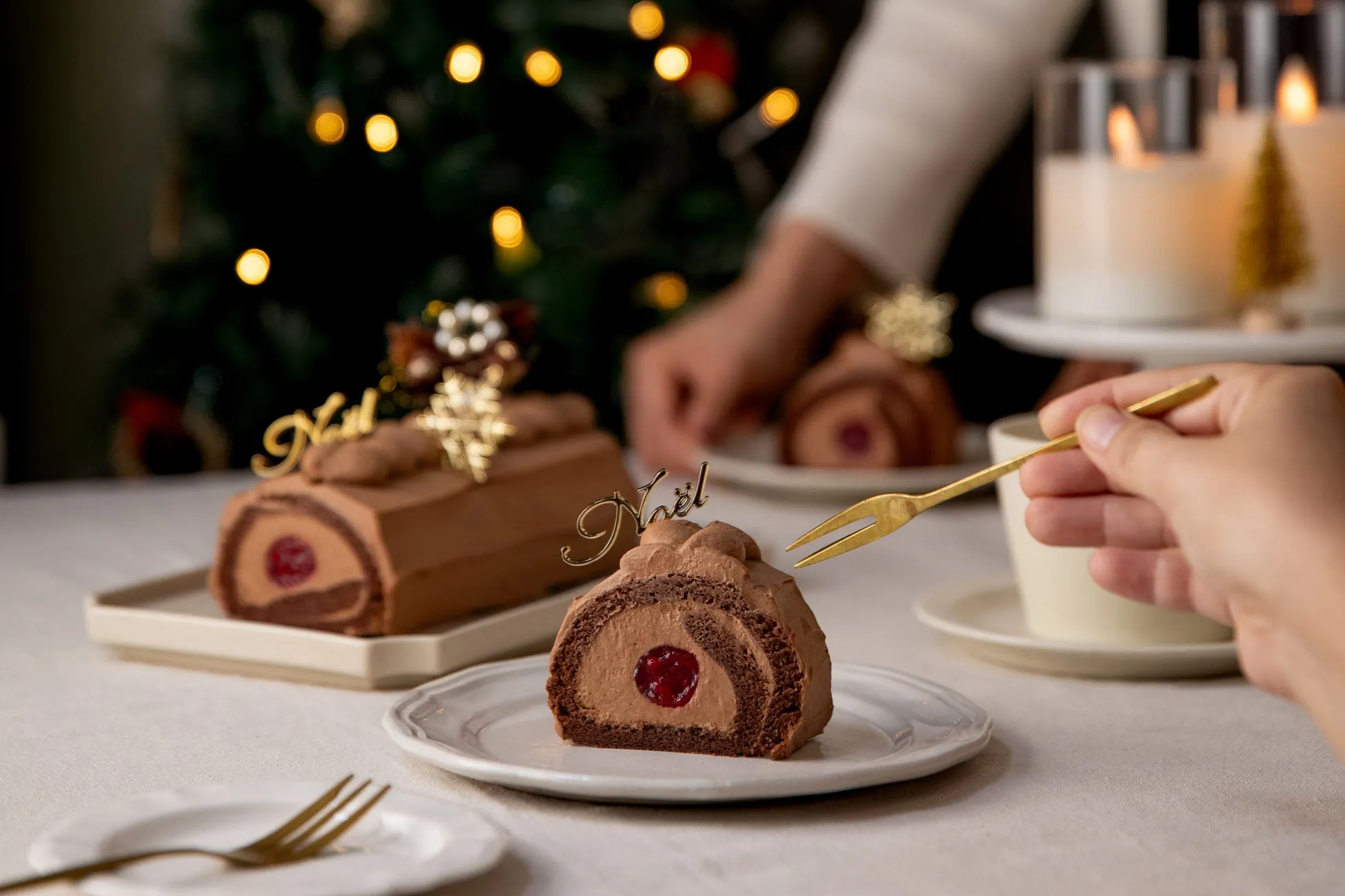 クリスマスをテーマにしたチョコレートケーキの写真。ケーキは、チョコレートクリームで覆われ、赤いチェリーが中心に入っています。背景には、クリスマスツリー、キャンドル、食器が見えます。