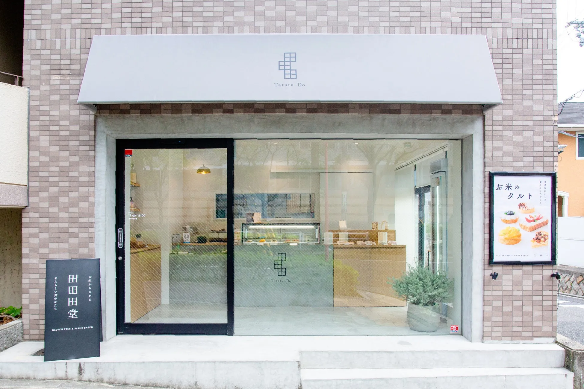 お店の外観。白を基調としたスタイリッシュな外観で、ガラス張りの窓からは店内の様子が伺える。看板には「田田堂」の文字と、グルテンフリー＆プラントベースのタルトの写真が掲載されている。