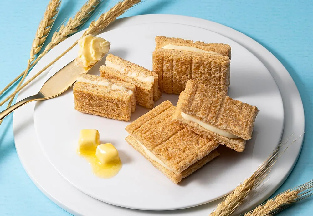 白い皿の上に、クリームを挟んだ四角いクッキーがいくつか並べられています。バターとバターナイフも置かれており、背景には麦の穂と水色の背景があります。