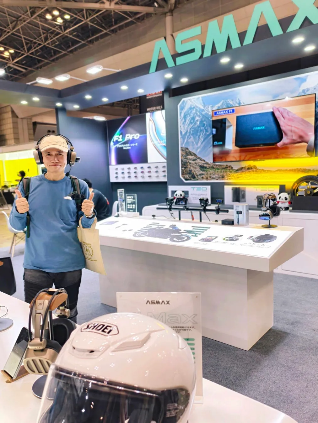 展示会で、ASMAXのブースでサムズアップをしている男性。