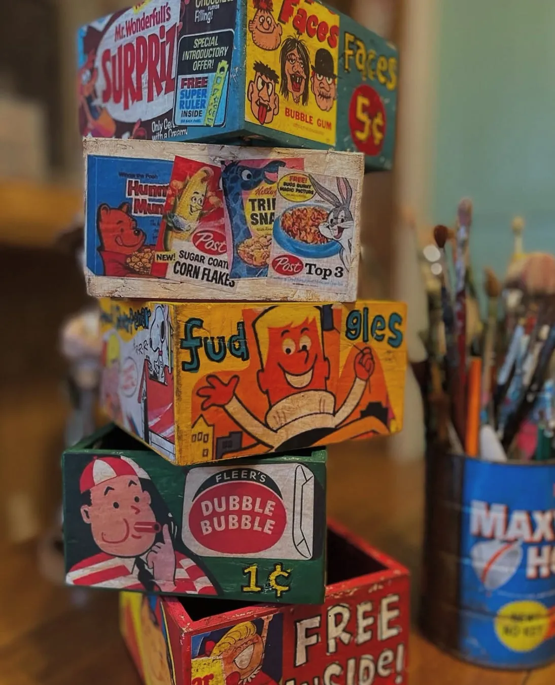 様々なお菓子のパッケージが描かれた木製の箱が積み重なっており、その横には絵筆立てが置かれている様子。
