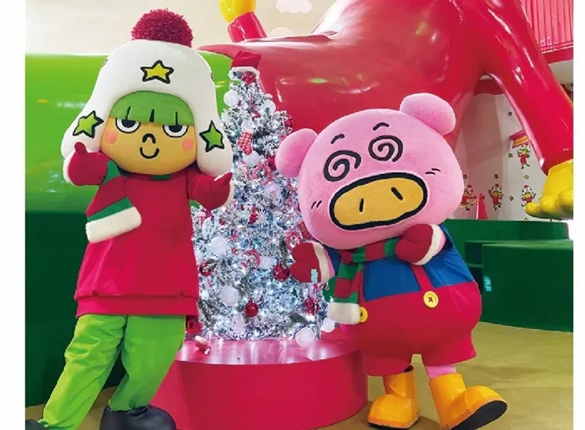 クリスマスをテーマにした、2体のゆるキャラが写っている写真です。左側のキャラクターは緑色の髪と服、赤い服を着ています。右側のキャラクターはピンク色の豚のキャラクターで、目が回っているように見えます。背景にはクリスマスツリーと大きな赤いオブジェがあります。