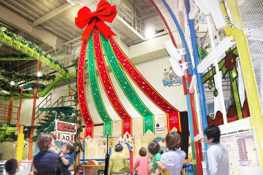 クリスマス装飾が施された子供向けの屋内遊び場の写真。天井から吊り下げられた大きな赤いリボンと、赤と緑のストライプのリボンが特徴的。