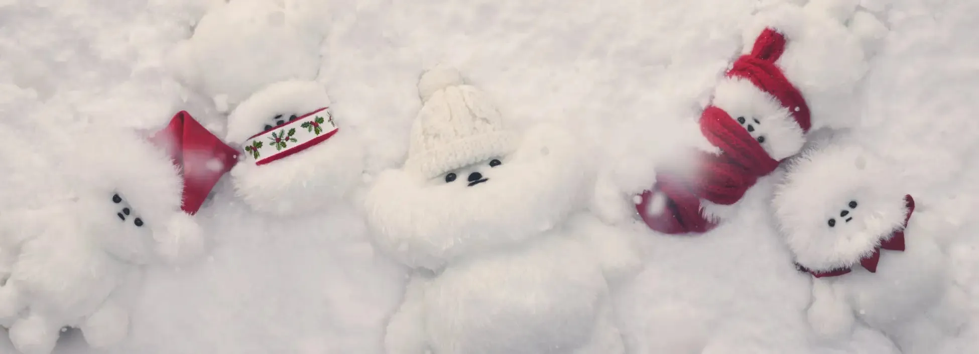 雪の中に横たわる、白い毛並みの犬のぬいぐるみ。赤いマフラーや帽子、飾りを身につけており、クリスマスを連想させる。