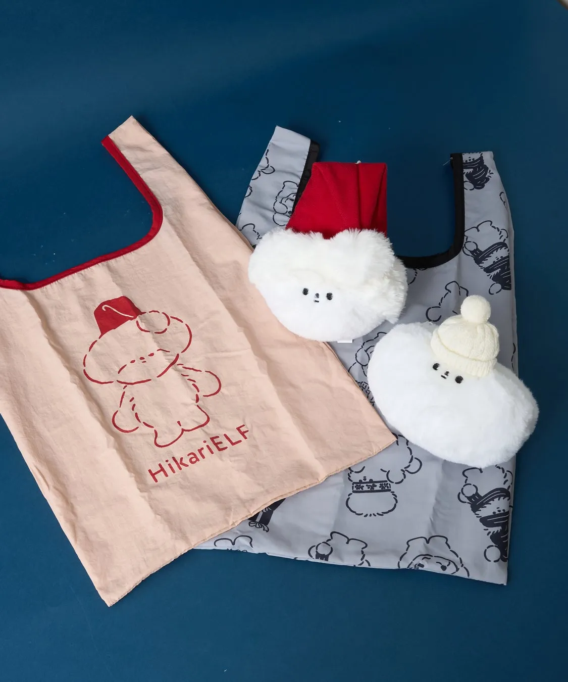 3つの異なるバッグが、青い背景に置かれています。ピンク色のバッグにはクマのイラストと「HikariELF」の文字があり、グレーのバッグには雲の模様と、白い雲の形をしたぬいぐるみと赤い帽子が飾られています。もう一つのバッグには、白い帽子をかぶった雲の形をしたぬいぐるみがあります。
