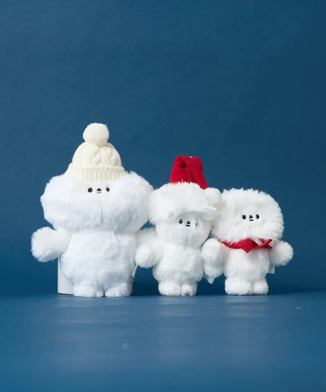 白いふわふわのぬいぐるみ3体。左のぬいぐるみは白いニット帽をかぶり、中央のぬいぐるみは赤い帽子をかぶり、右のぬいぐるみは赤いスカーフを巻いている。背景は青色。