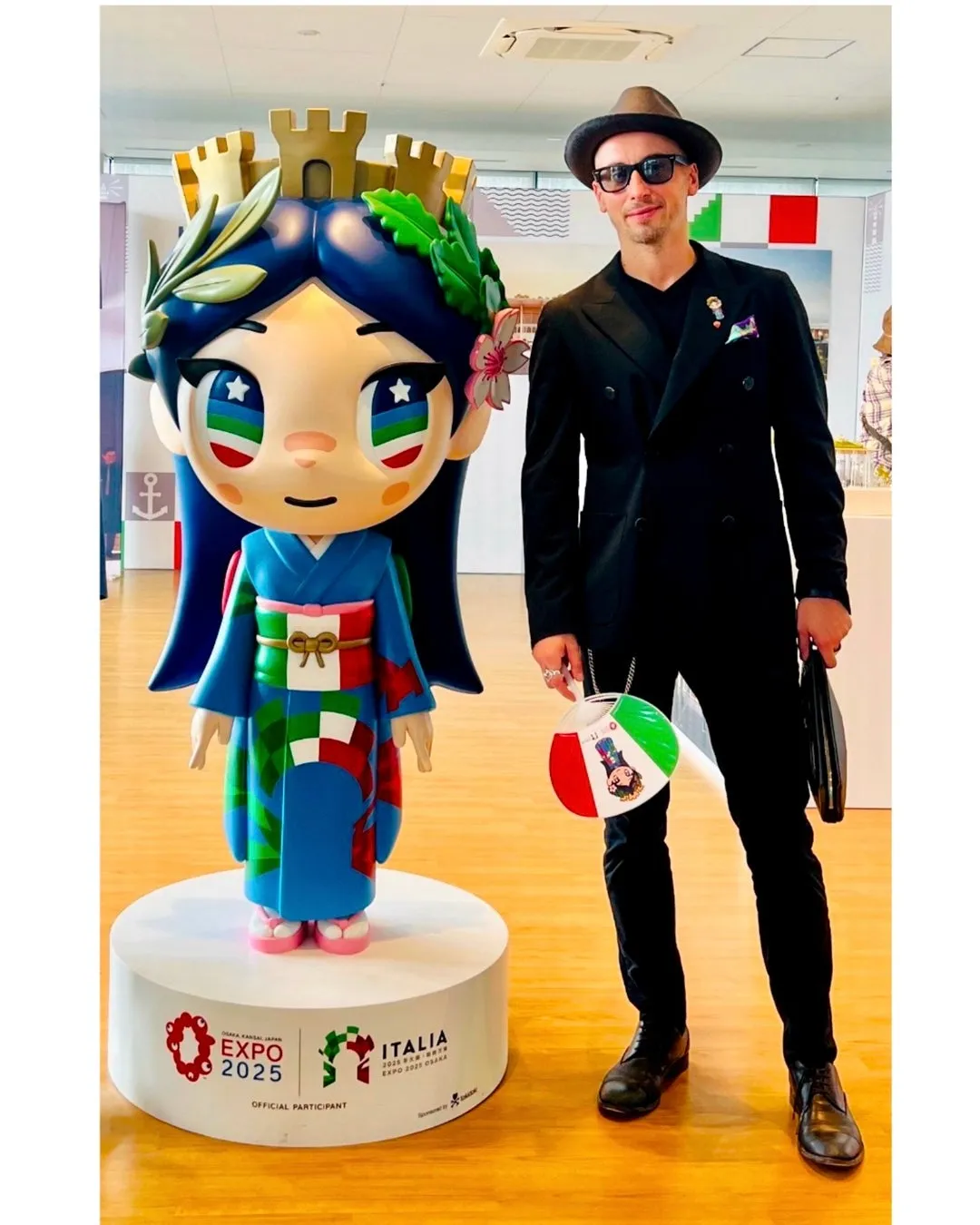 男性が、日本の万博のマスコットキャラクターとイタリアの国旗をモチーフにしたバッグを持って立っている様子。