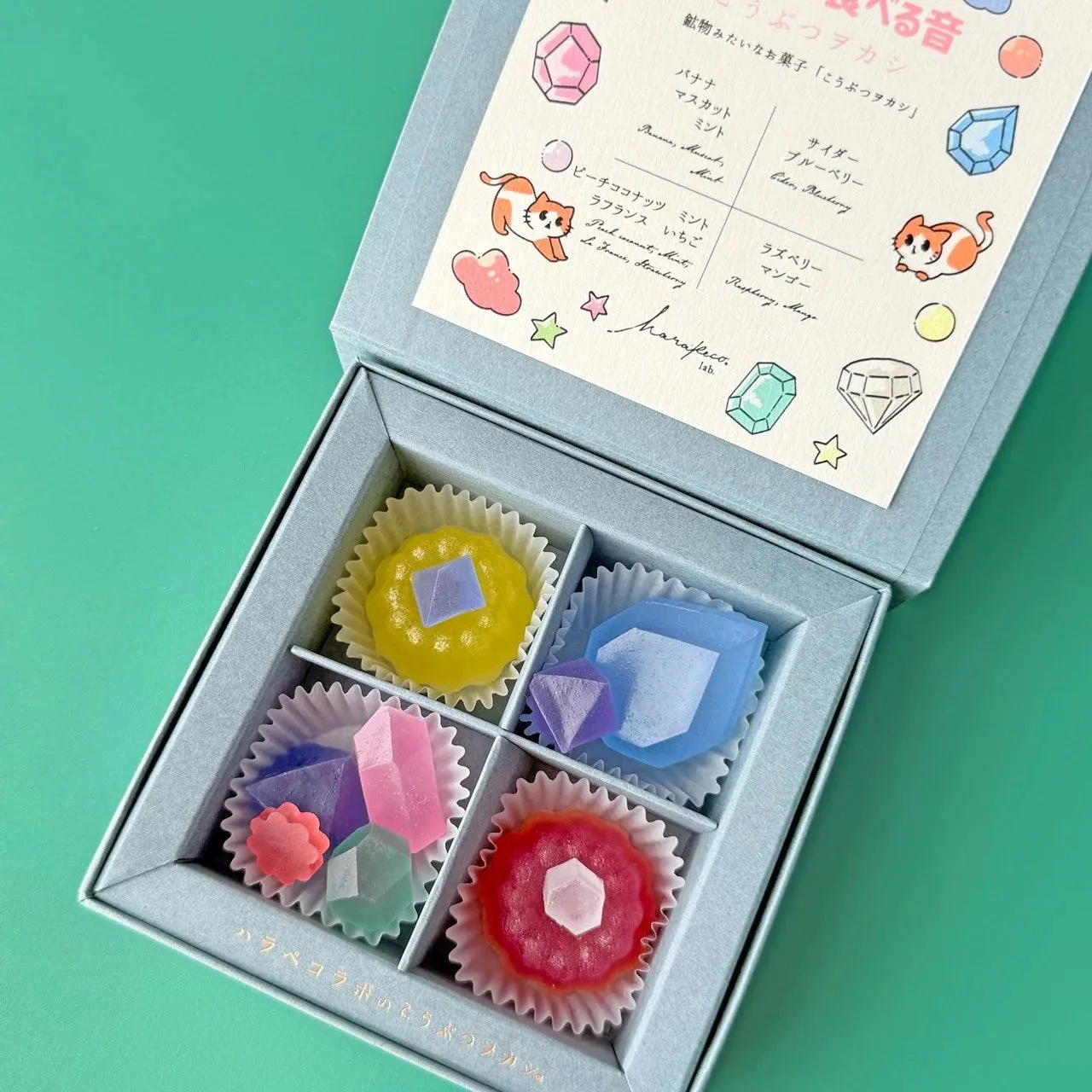 宝石のような形をしたカラフルなゼリー菓子が、ギフトボックスに入っている様子を写した写真です。箱の中には、様々な色と形のゼリー菓子が並べられており、それぞれの味や素材が記載されたカードも添えられています。全体的に可愛らしく、贈り物やプレゼントにも適した印象を与えます。