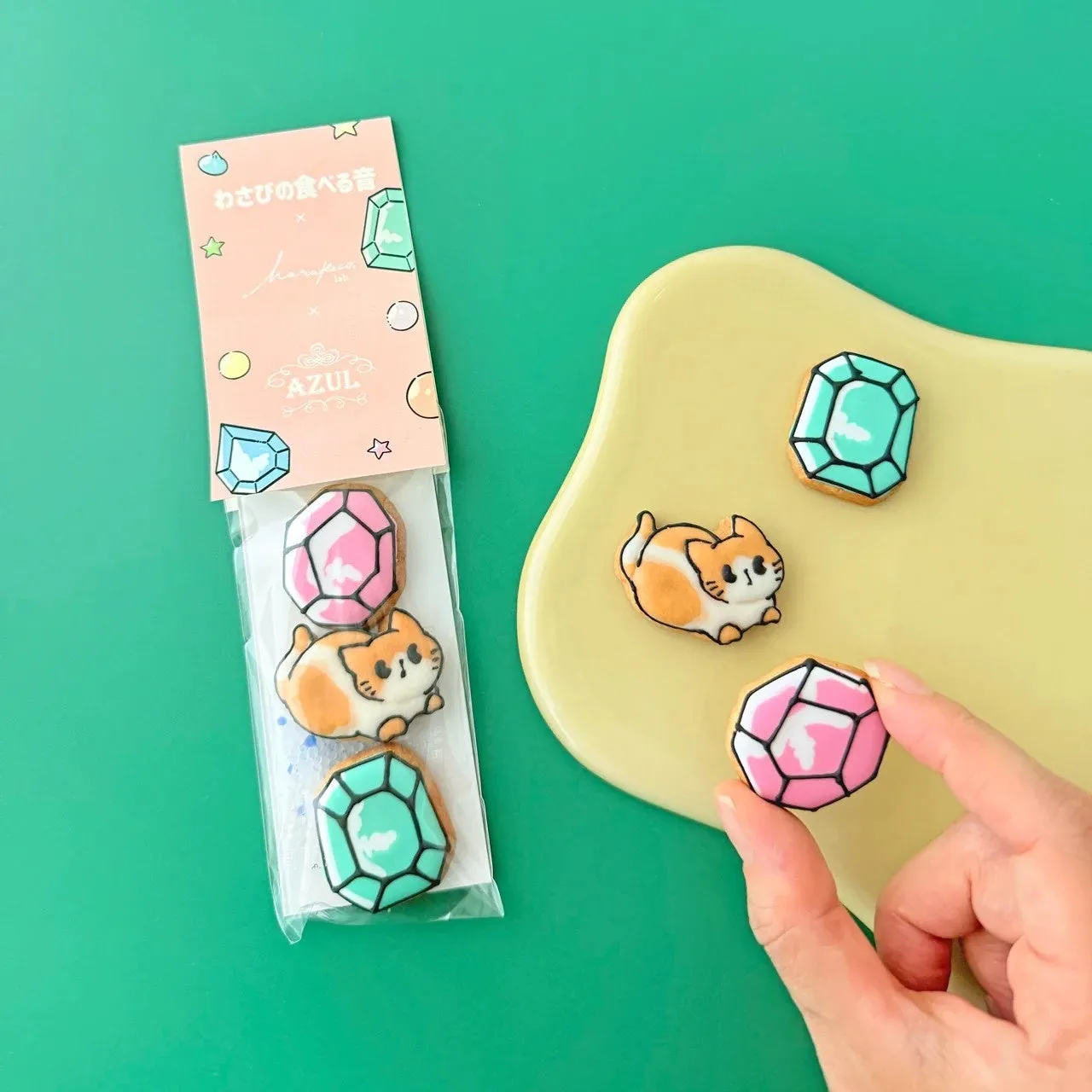 宝石と猫の形をしたアイシングクッキーが写った写真。宝石クッキーはピンクと緑の2種類、猫クッキーはオレンジと白。手前には宝石クッキーが手に持たれ、奥には黄色い皿にクッキーが並んでいる。背景は緑色。