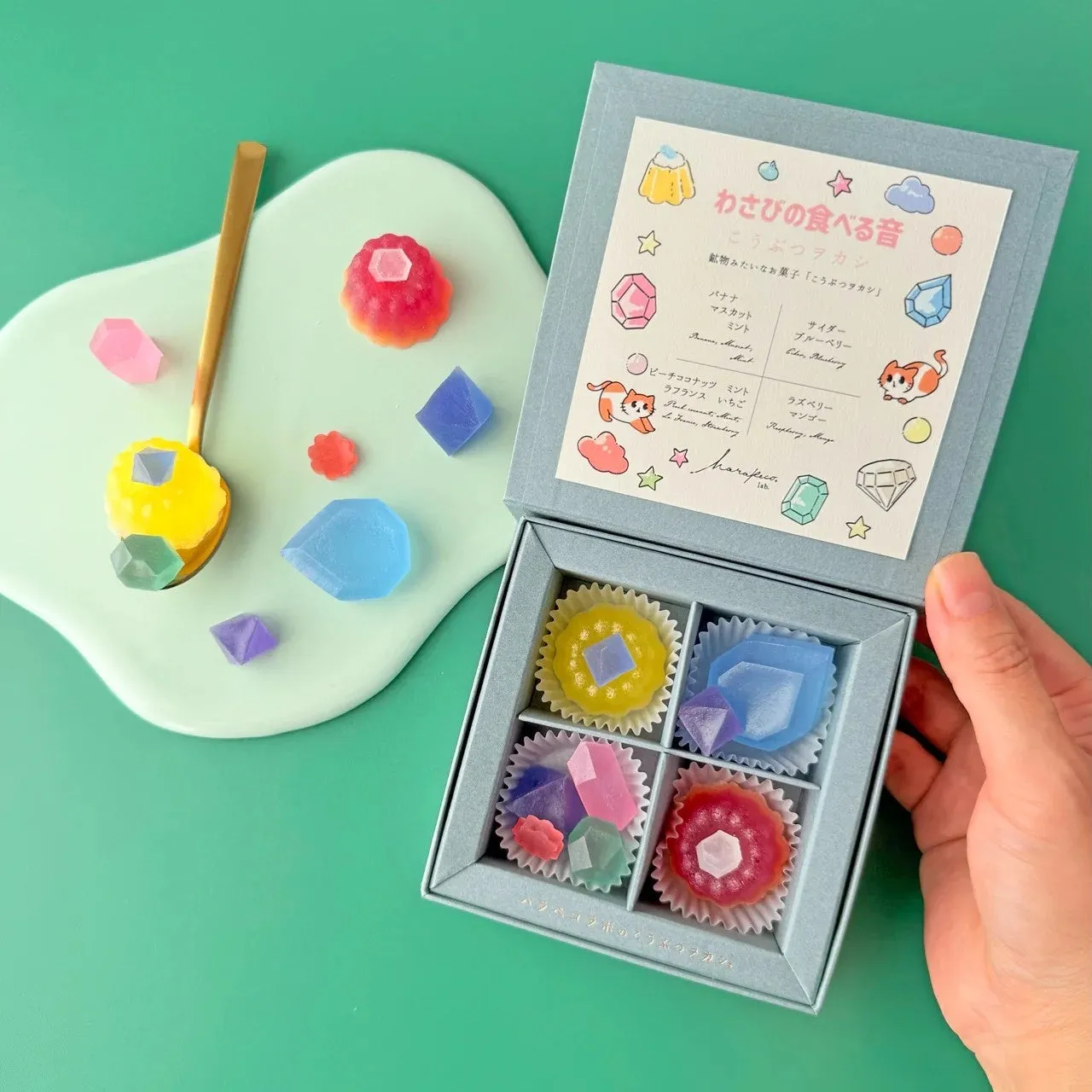 宝石のような美しい見た目の和菓子が詰め合わせになった商品の写真。淡いブルーのギフトボックスの中に、様々な色の宝石を模した美しい寒天菓子が並んでいる。スプーンに乗せられたものや、ギフトボックスの外に散りばめられたものも。