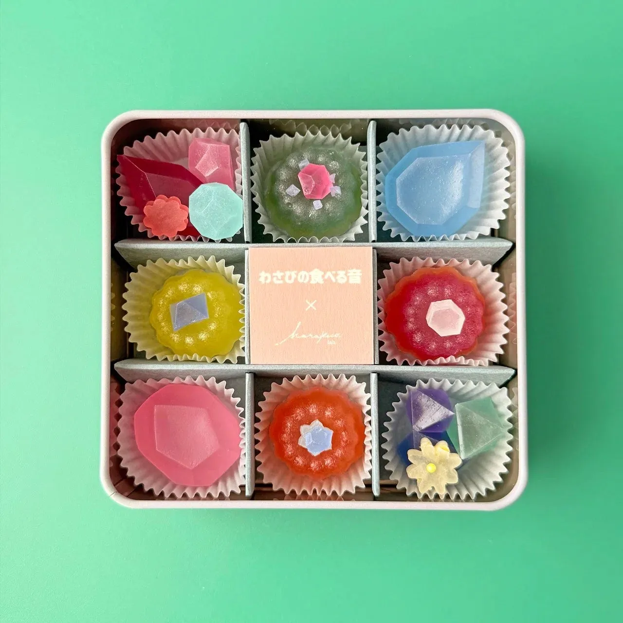 宝石のような見た目のカラフルな和菓子が9つ入ったギフトボックス。それぞれの和菓子は異なる色と形で、まるで宝石のよう。中央には「わさびの食べる音」と書かれたカードが配置されている。