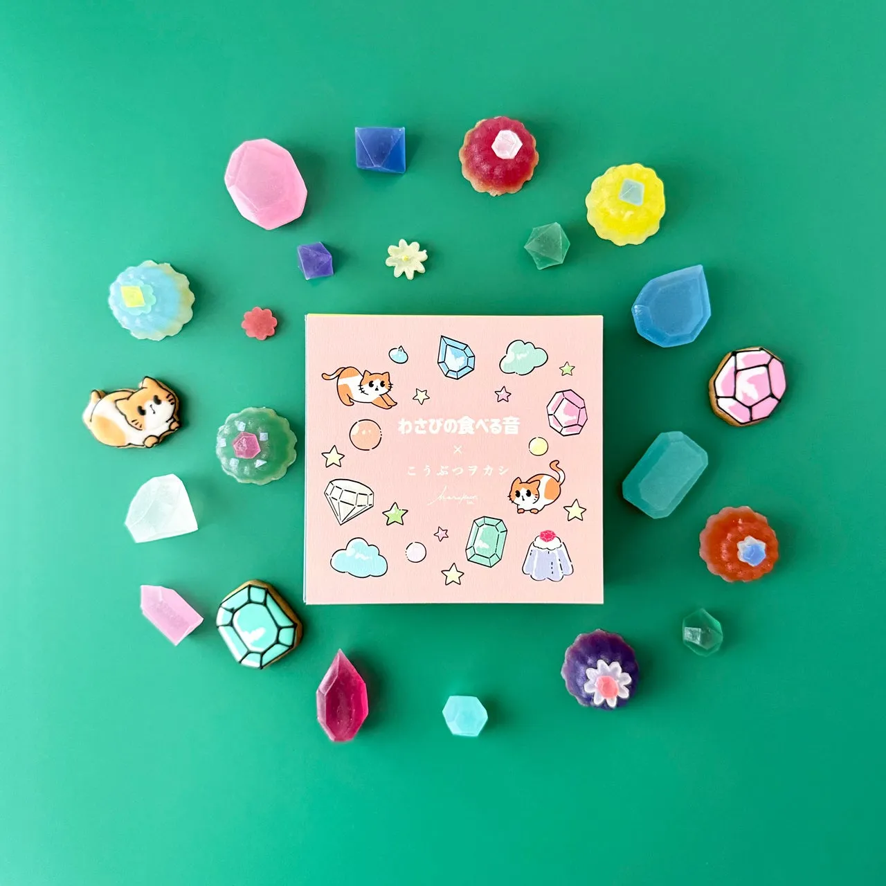 宝石のような美しい和菓子が、緑色の背景に配置された写真。中央には、和菓子が詰められた箱があり、周りには様々な形の和菓子が散りばめられています。全体的に、可愛らしくて幻想的な印象を与える写真です。