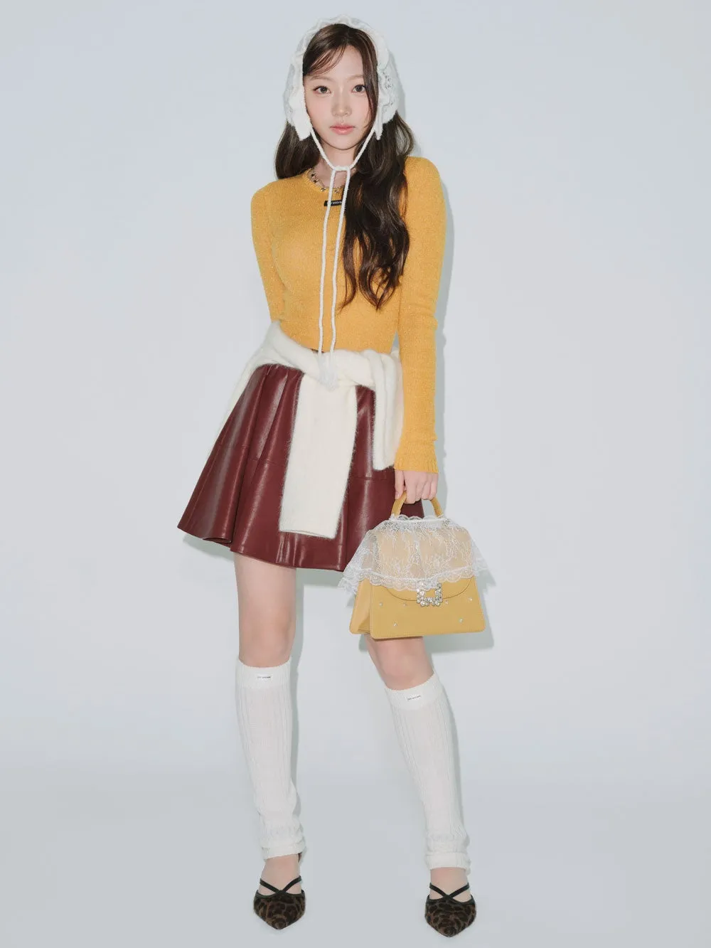女性が白い背景でポーズをとっています。彼女は、黄色いセーター、革のスカート、白いストール、白いソックス、ヒョウ柄の靴を履いています。
