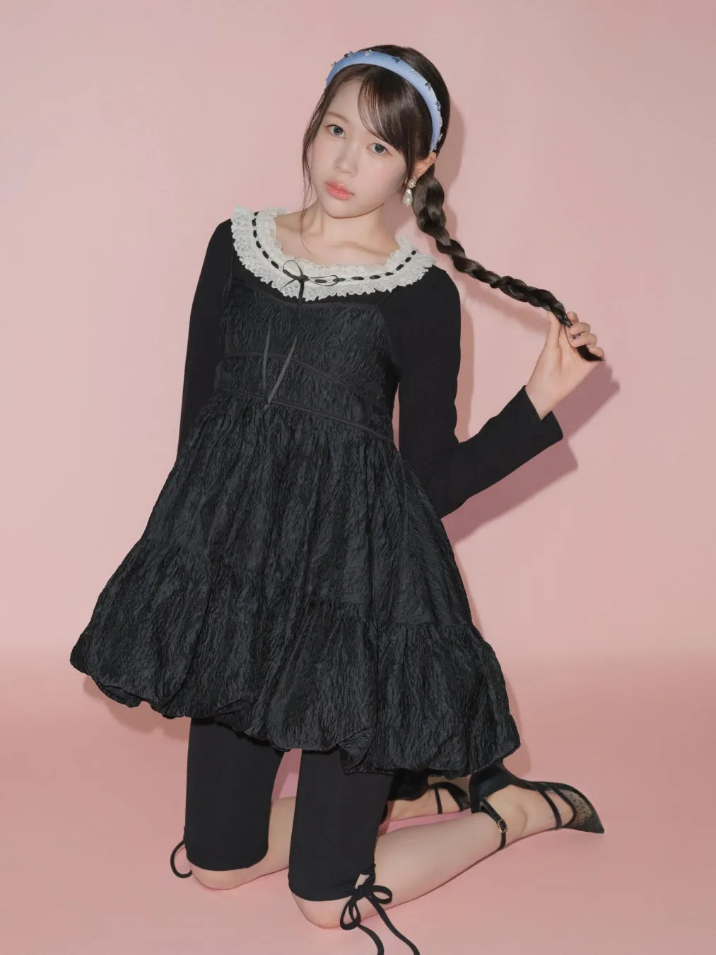 ピンク色の背景に、黒いドレスを着て三つ編みをした若い女性が膝立ちしています。