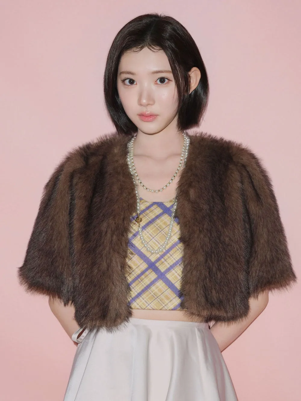 ピンク色の背景の前に立つ女性のポートレート。彼女は短い黒髪で、茶色の毛皮のジャケット、チェック柄のトップス、白いスカートを着ている。