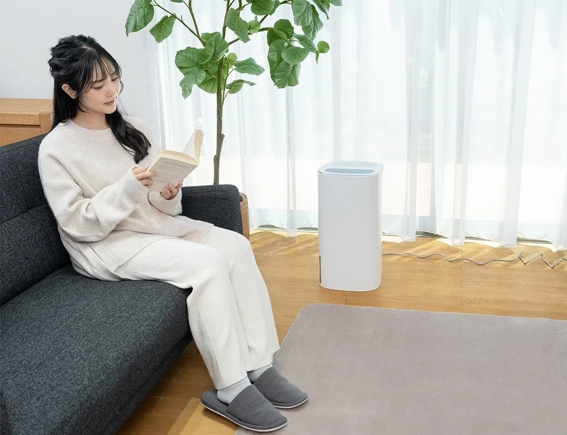 女性がソファーに座り、本を読んでいる様子。白い加湿器がそばに置かれ、カーテンと観葉植物が背景にある。