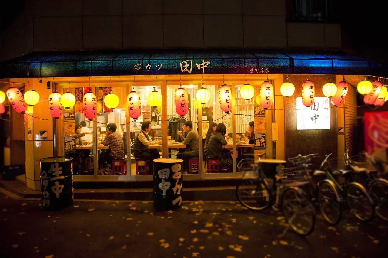 日本の居酒屋の夜景。提灯が飾られ、店内には人々が食事を楽しんでいる様子が写っています。