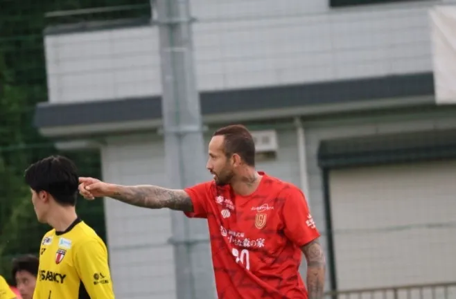 サッカーの試合中の写真。赤いユニフォームを着た選手が、指をさして指示を出している。近くには、黄色いユニフォームを着た別の選手がいる。