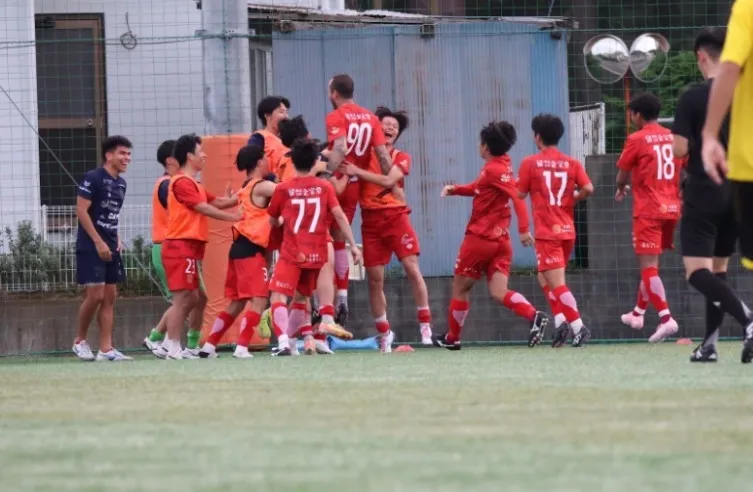 サッカーの試合で、赤いユニフォームを着た選手たちが勝利を祝って抱き合っている様子。