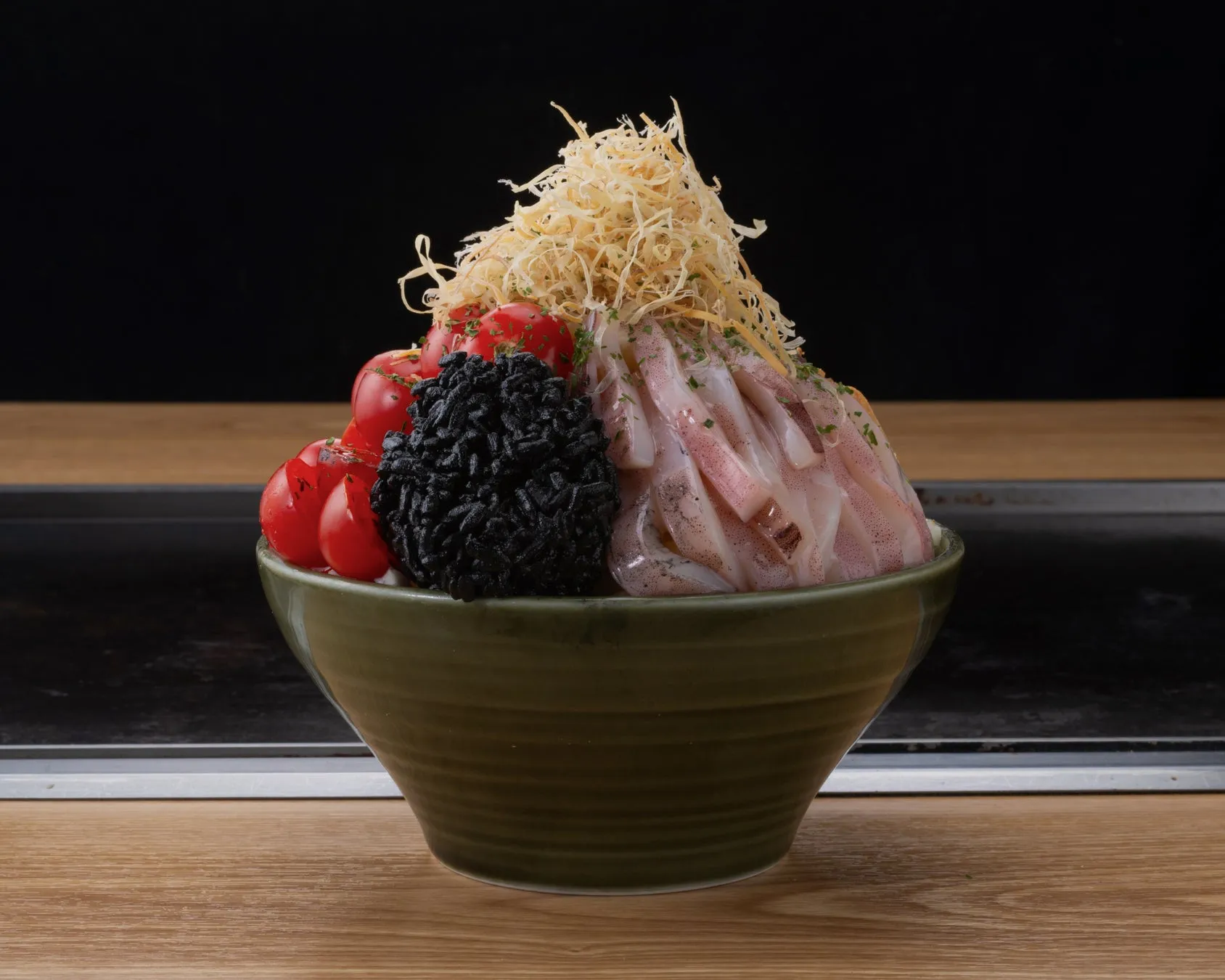 緑色のボウルに入った料理のクローズアップ写真。料理には、トマト、黒い食べ物、イカ、揚げた食べ物などが含まれています。料理は木製のテーブルの上に置かれています。
