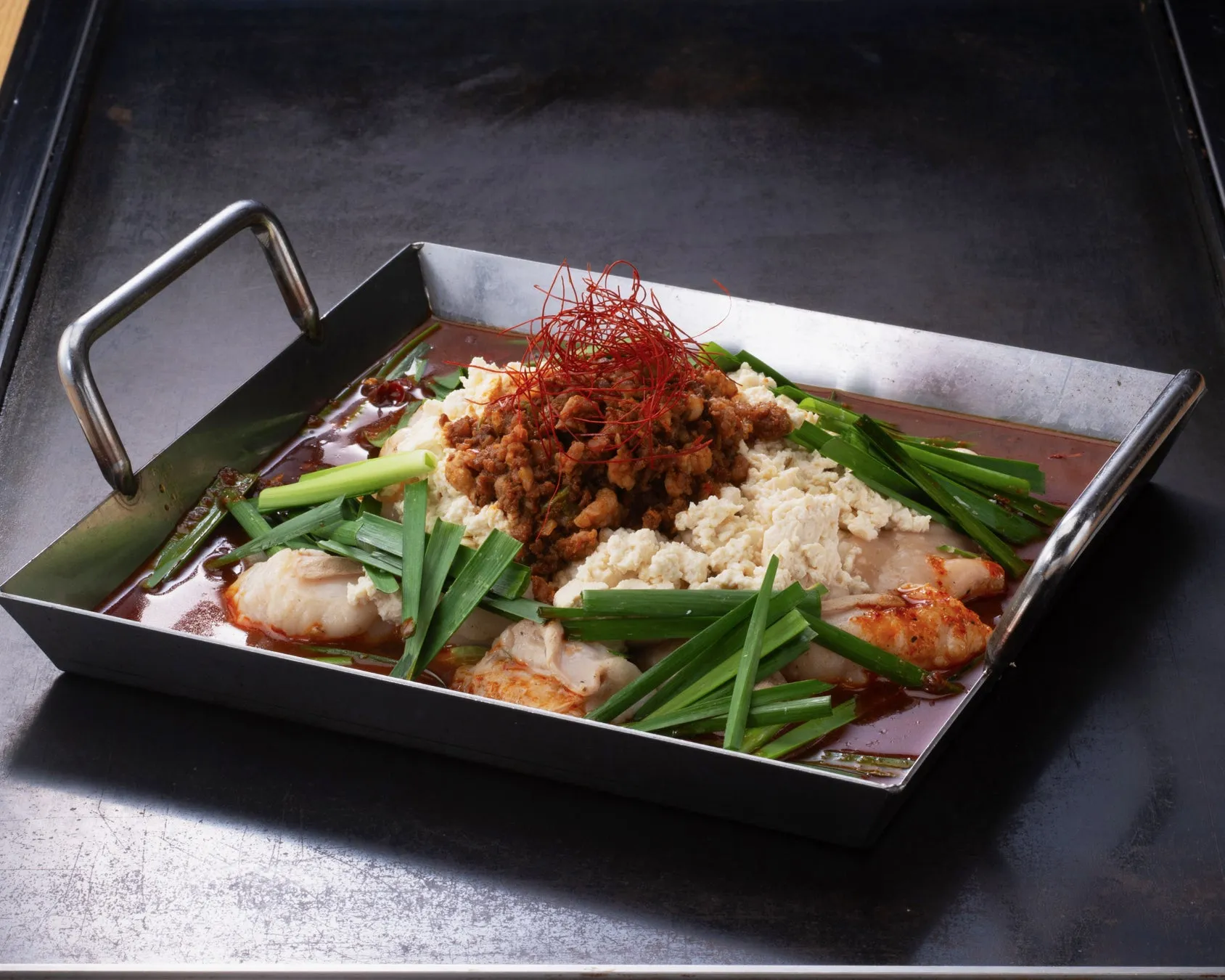 長方形の鉄板に盛られた料理の写真。中央にはひき肉のようなもの、その周りには豆腐、ニラが添えられ、唐辛子の糸がトッピングされている。