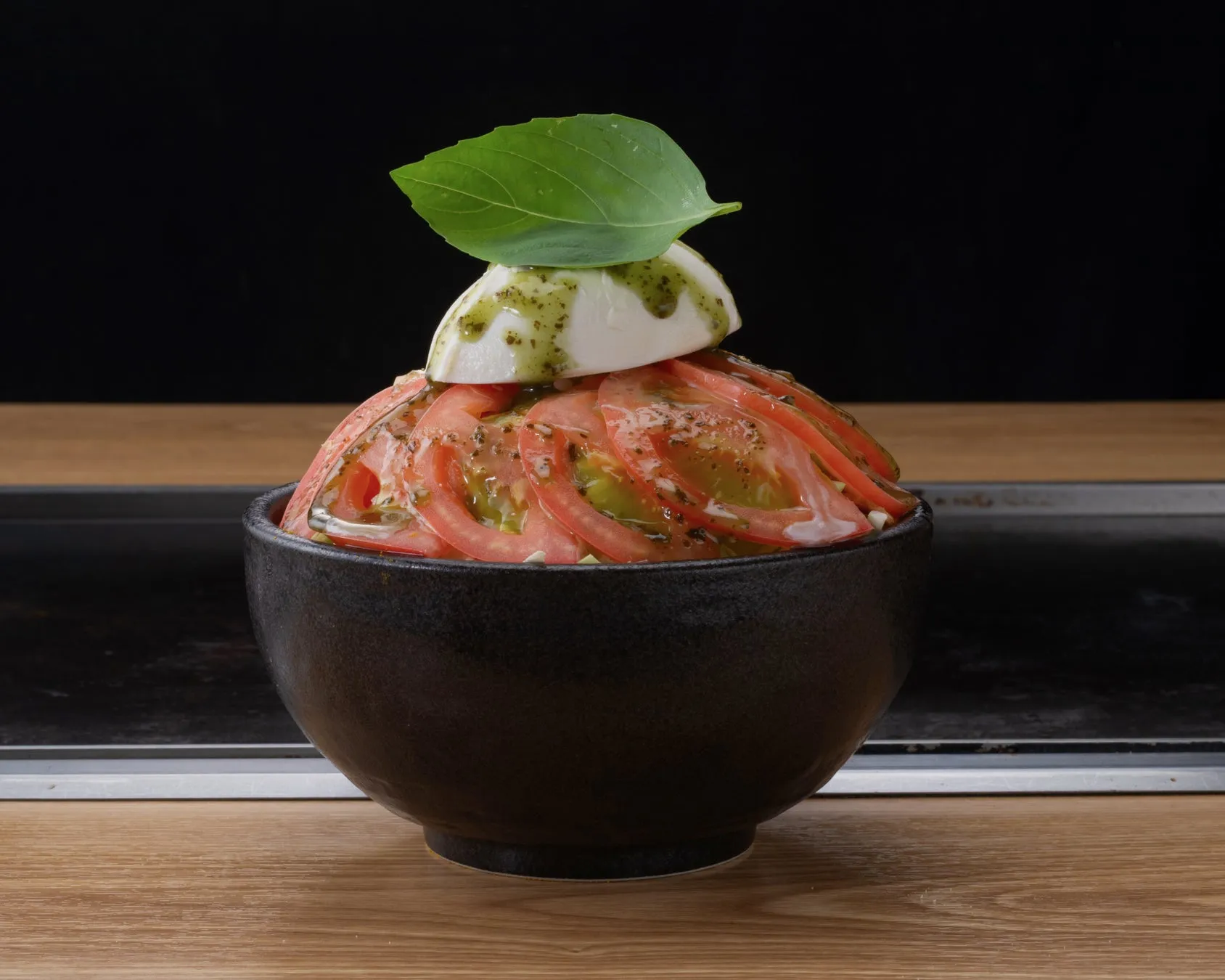 トマトとモッツァレラチーズを重ねて盛り付け、バジルを添えた料理のクローズアップ写真。黒い器に盛られており、木製のテーブルの上に置かれている。