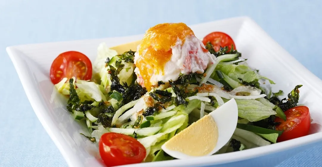 白い器に盛られたサラダ。サラダには、レタス、キュウリ、トマト、海苔、ゆで卵、カニカマ、ソースがトッピングされている。