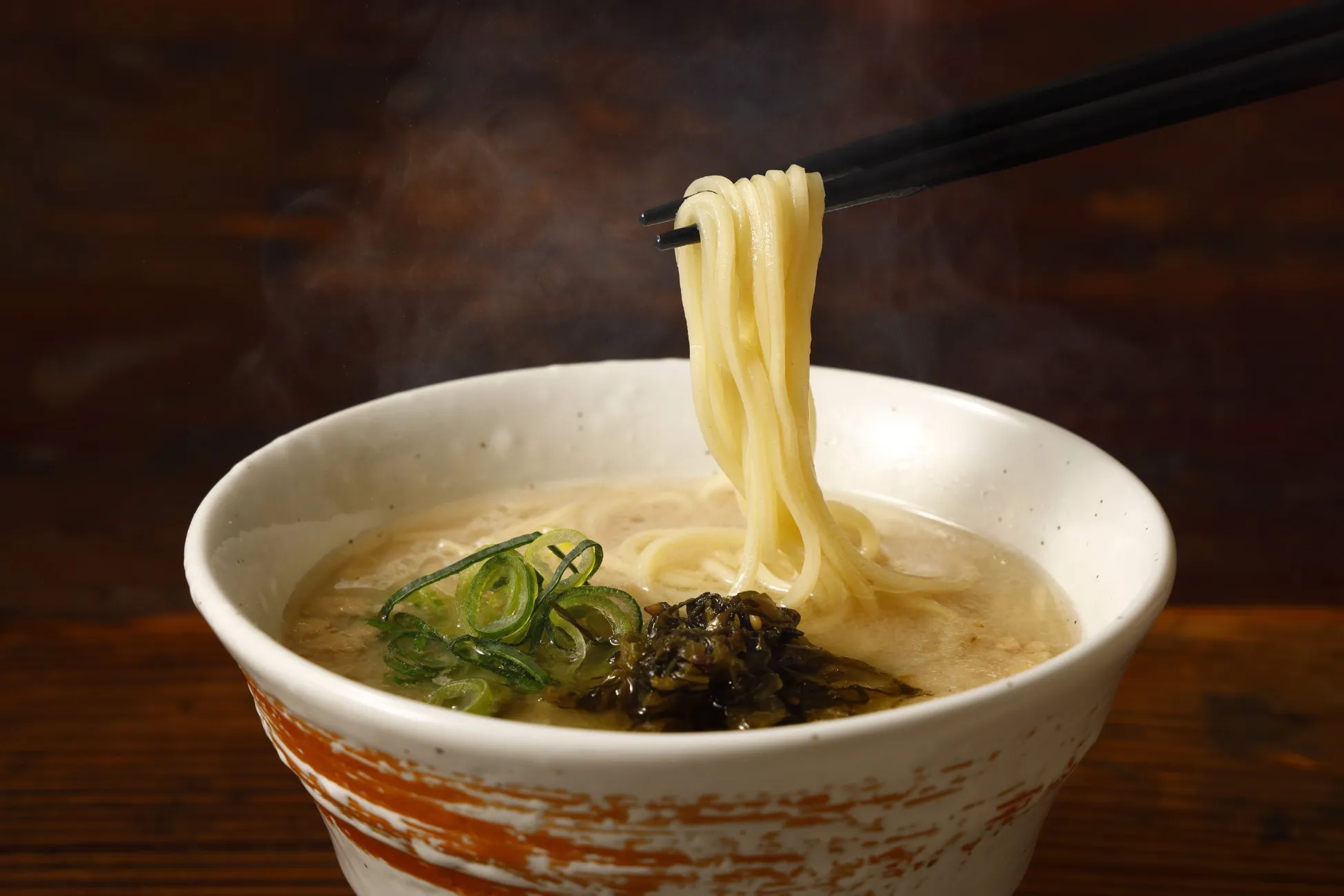 湯気が立ち上るラーメン。箸で持ち上げられた麺、ネギや漬物がトッピングされたスープが特徴的。