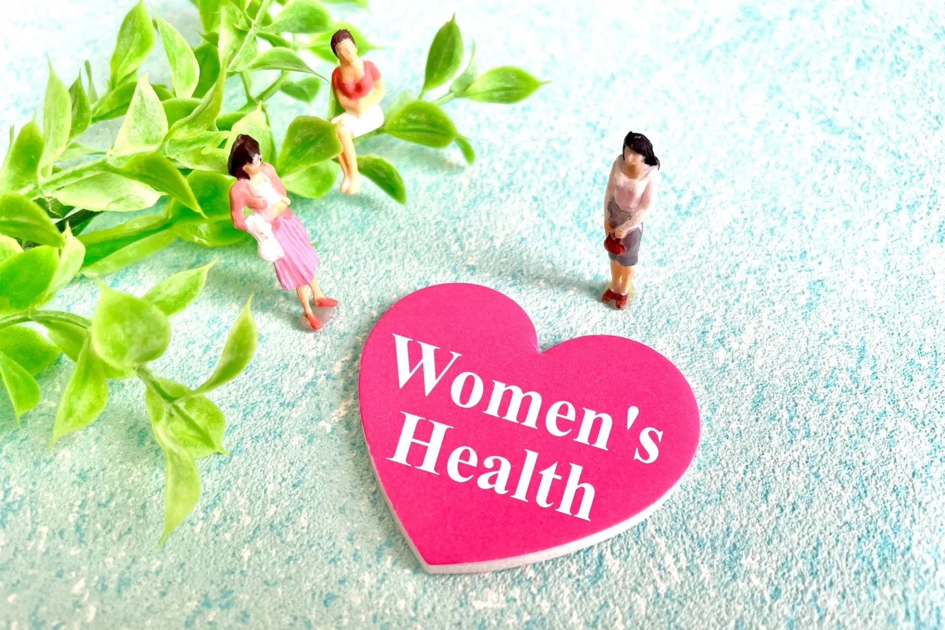 ピンク色のハート型のプレートに「Women's Health」の文字が書かれており、3人の女性のミニチュアが置かれた画像です。背景には緑の葉が飾られています。