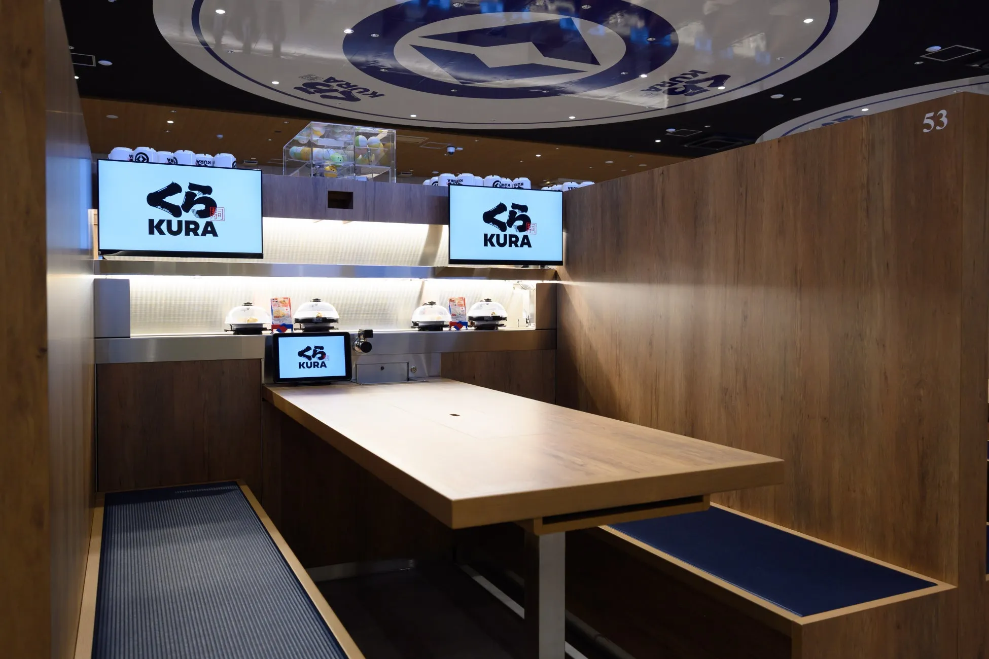 木目調の内装が印象的な、くら寿司の店舗内の写真です。テーブル席と、カウンター席のような席があります。天井にはお店のロゴが描かれています。