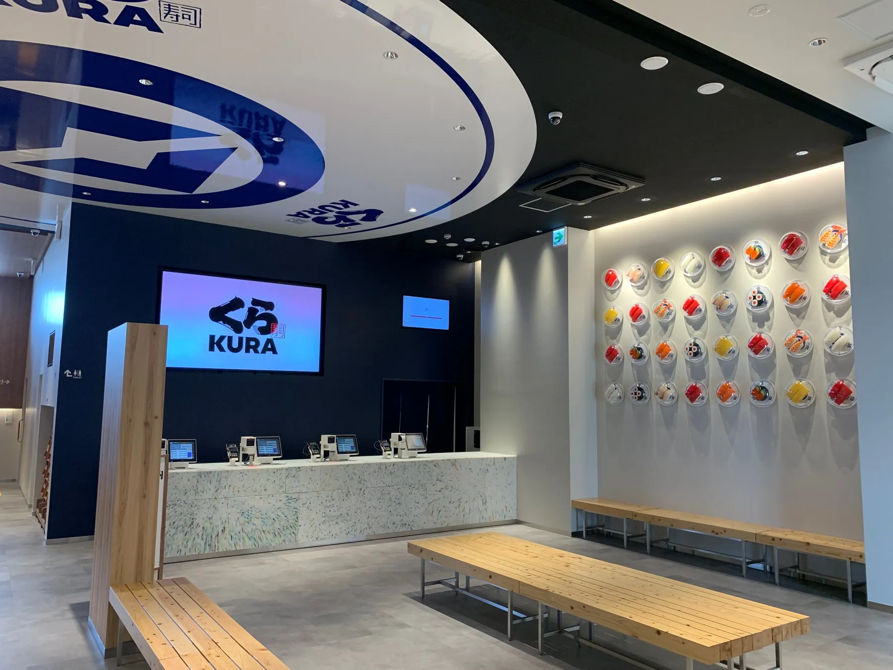 くら寿司の店内の写真。壁には寿司の見本が飾られており、天井にはロゴがデザインされています。