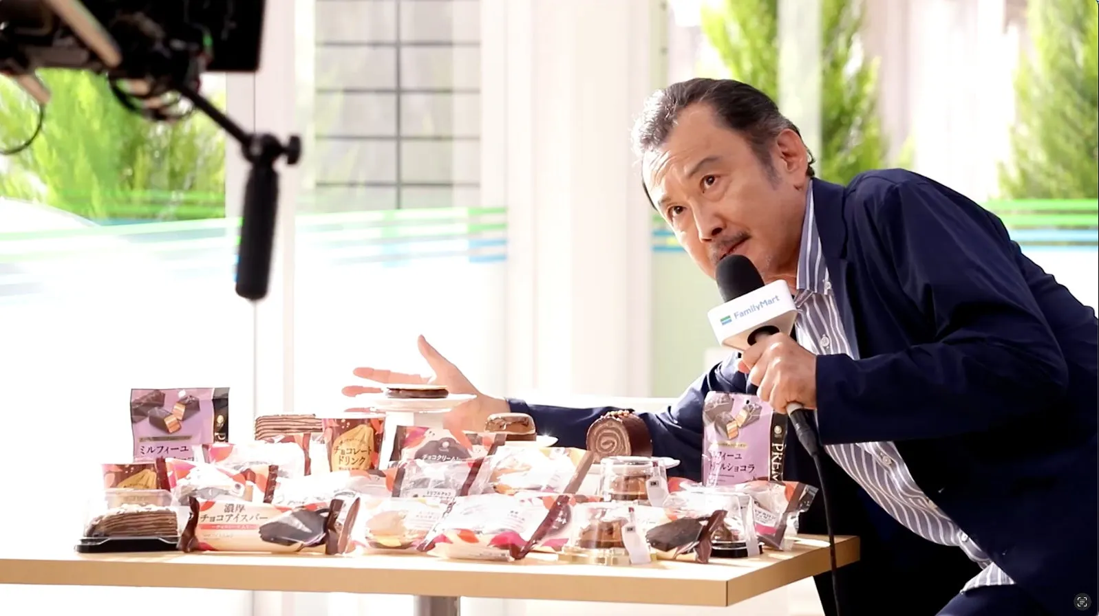 女性がFamilyMartの商品の前でマイクを持って話している様子。テーブルには様々なスイーツが並んでいる。