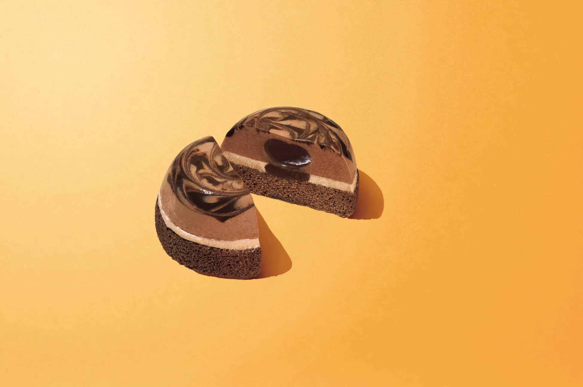 オレンジ色の背景に、カットされたチョコレートケーキが2つ配置されています。ケーキは、チョコレートスポンジケーキ、チョコレートムース、チョコレートゼリーの層で構成されています。上部にはチョコレートのマーブル模様があり、カットされた部分から各層が見えています。