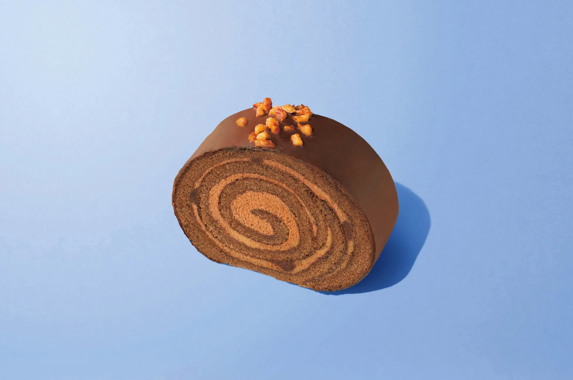 青色の背景に、チョコレートロールケーキが一つ置かれています。ロールケーキは、チョコレートでコーティングされ、上にナッツがトッピングされています。ケーキの断面には、渦巻き模様が見えます。