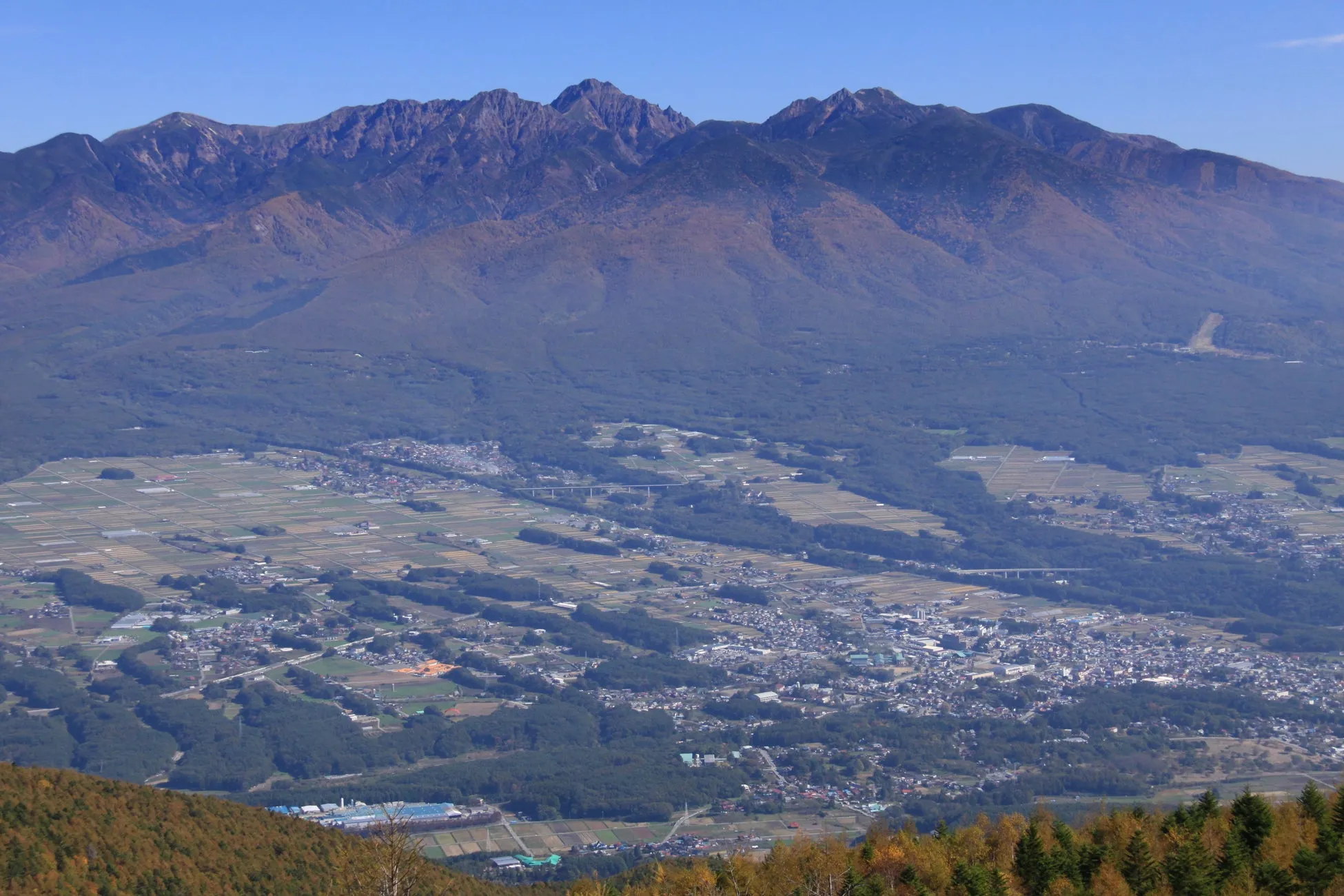 山々と街並みの広角写真。遠くには山々がそびえ立ち、その手前には広大な平野と街並みが広がっている。鮮やかな青空の下、自然と都市が調和した美しい風景。