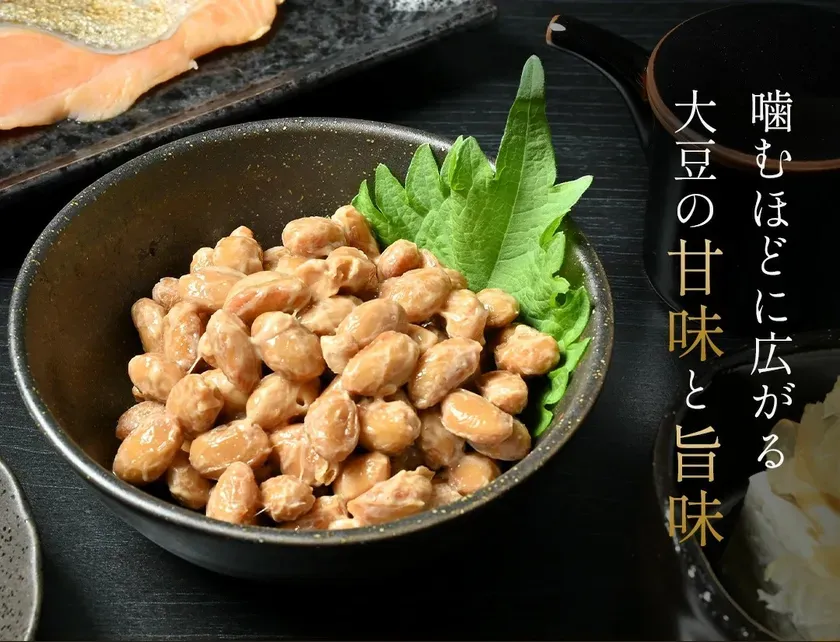 納豆のクローズアップ写真。納豆は黒い器に入っており、緑の葉で飾られている。背景には、サーモンと薬味のようなものが見える。