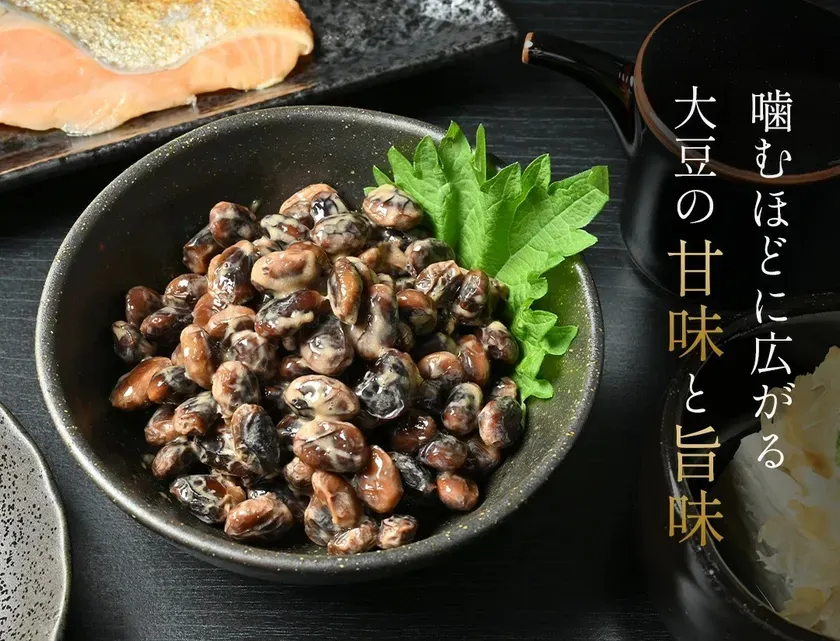 納豆のクローズアップ写真。納豆は黒い器に入っており、緑の葉で飾られている。背景には、サーモンと薬味のようなものが見える。