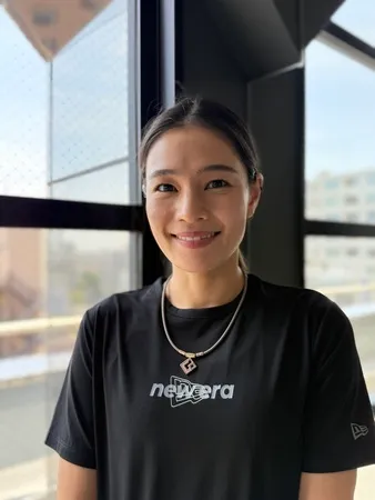 窓際に立つ女性。黒いTシャツを着て、笑顔でカメラを見つめている。彼女はネックレスをしており、背後には明るい窓と街並みが見える。