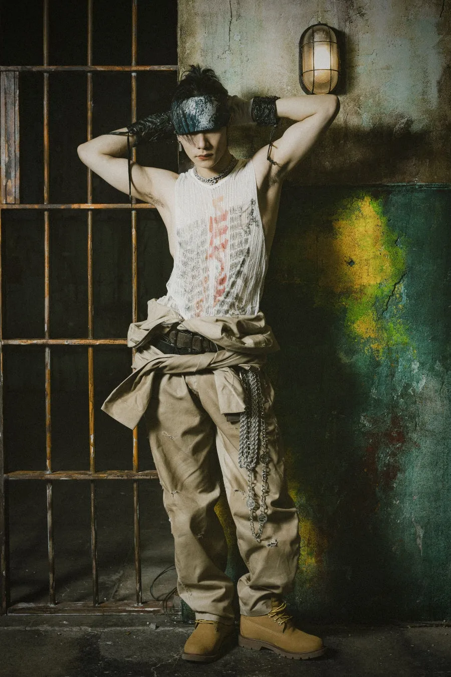 男性が刑務所の壁に寄りかかってポーズをとっている写真。彼は白いタンクトップとカーキ色のパンツを着用し、頭にはバンダナを巻いています。背景には刑務所の鉄格子と壁に掛けられたライトがあります。