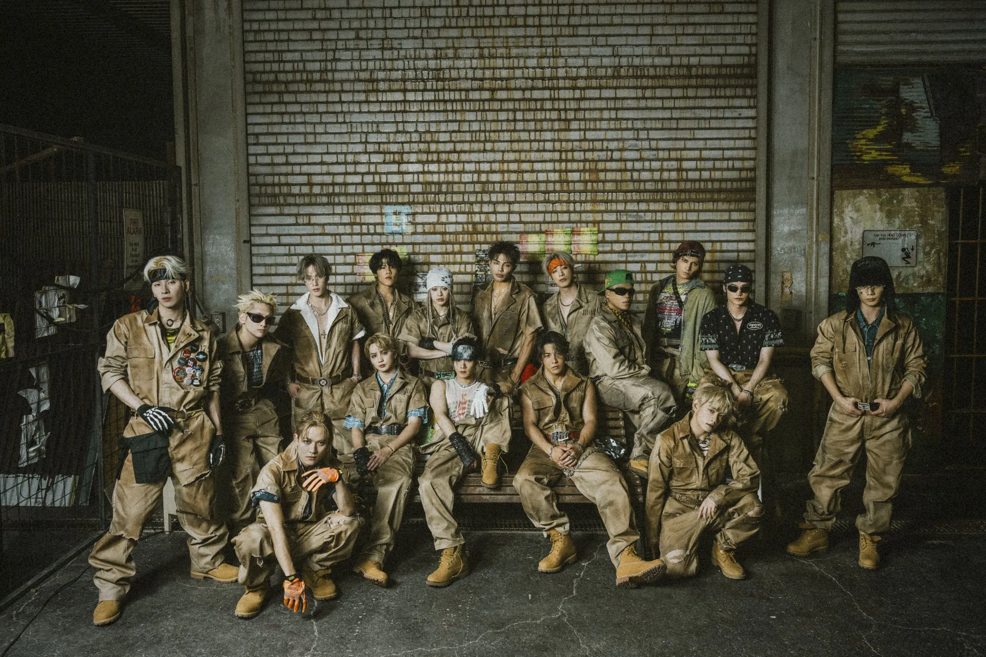 茶色のオーバーオールを着た17人の男性グループが、レンガの壁の前でポーズをとっています。彼らは、様々な髪型やアクセサリーを身につけています。彼らは、建物の外にいるように見えます。