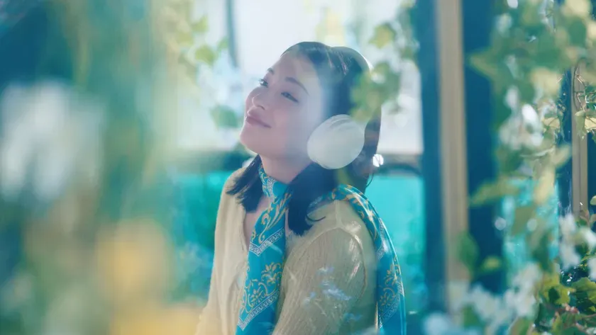 女性がヘッドホンをして目を閉じている様子。背景には緑の葉や水色の背景が見える。