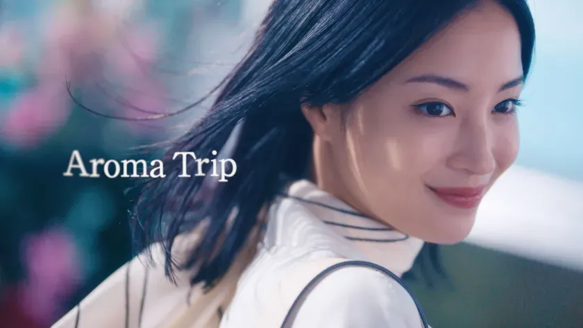 女性のポートレート。長い黒髪で、風になびいています。彼女は微笑んでいて、カメラのほうを見ています。背景には、ぼやけた花と青い空があります。