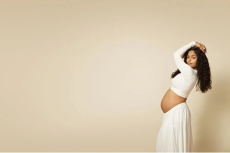 妊娠中の女性が白い服を着て、手を頭の上に置いてポーズをとっています。