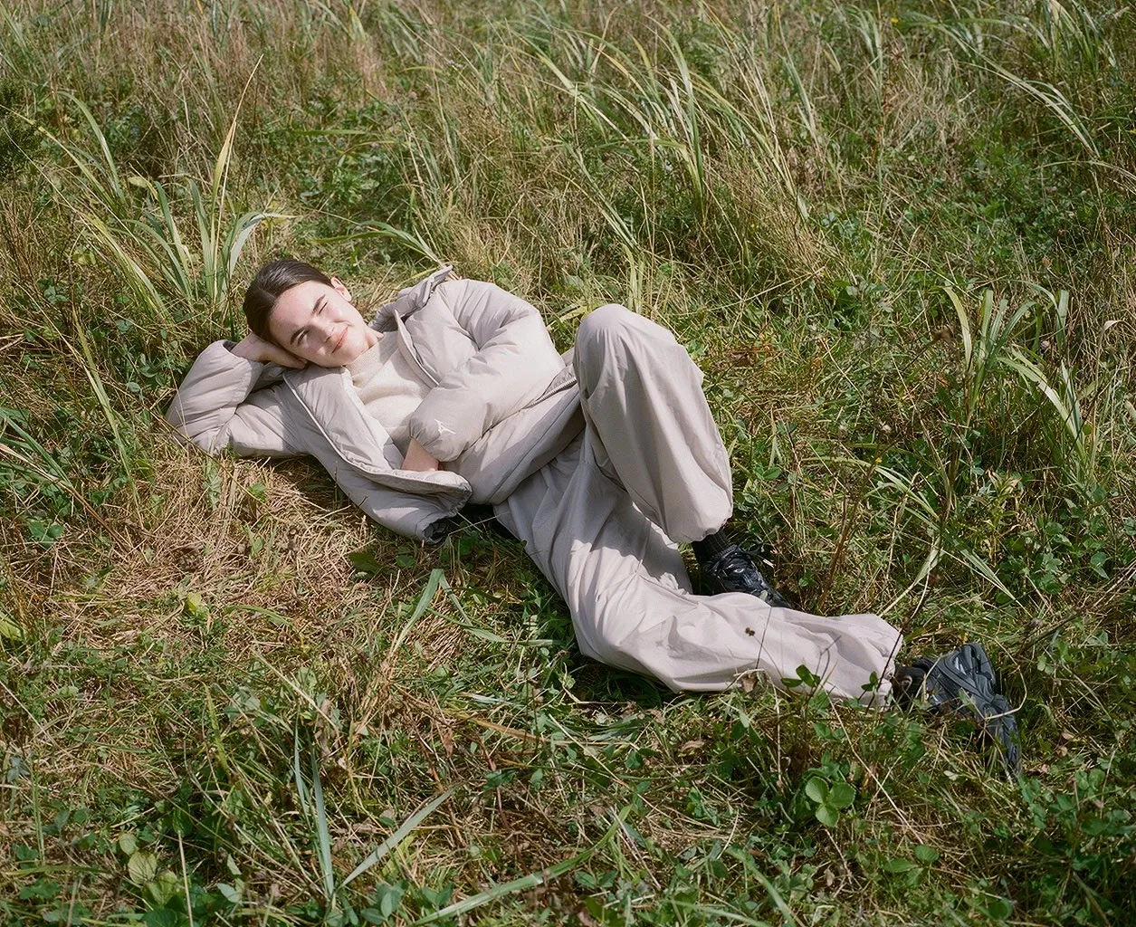 野原でくつろぐ女性。ベージュのダウンジャケットとパンツを着て、草の上に横たわっている。