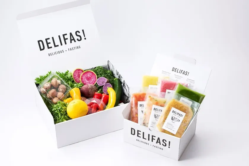 2つの白い箱に入った食べ物の画像。左の箱には野菜が、右の箱にはパックされた食べ物が入っています。どちらの箱にも「DELIFAS!」のロゴと「DELICIOUS + FASTING」という文字が書かれています。