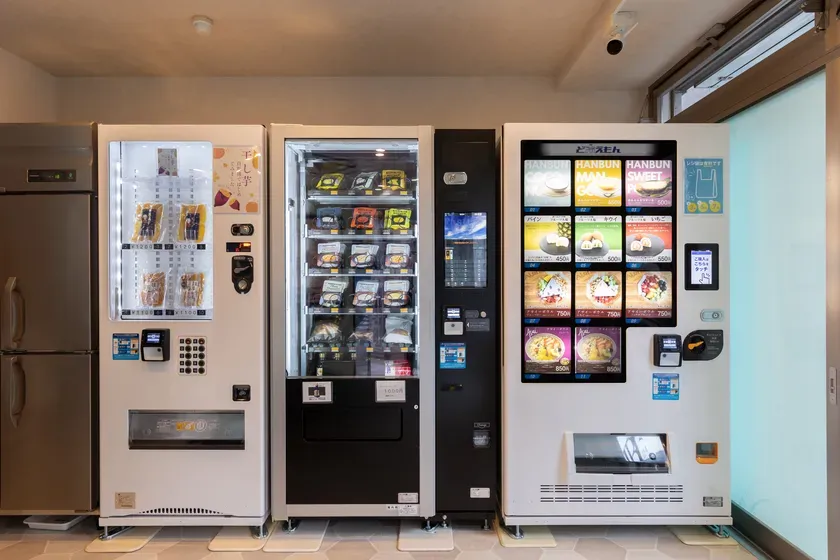 自動販売機が並んだ写真。様々な商品が販売されており、メニューや価格が表示されている。店内には監視カメラも設置されている。