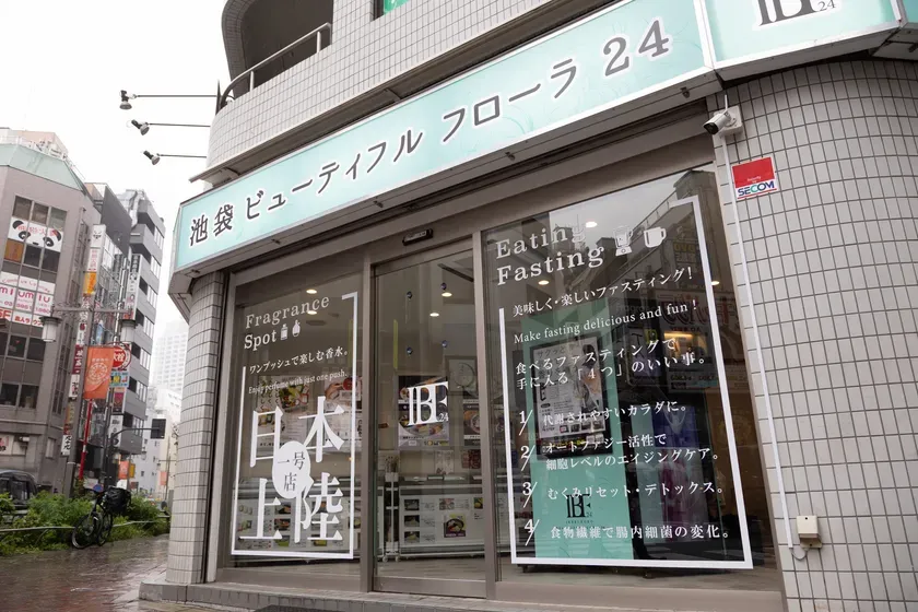池袋にある、ビューティフルフローラ24というお店の外観の写真です。お店の看板には日本語で店名が書かれており、窓には「Eating Fasting」や「Fragrance Spot」といった文字が見えます。お店の前には自転車が止まっており、雨が降っているようです。