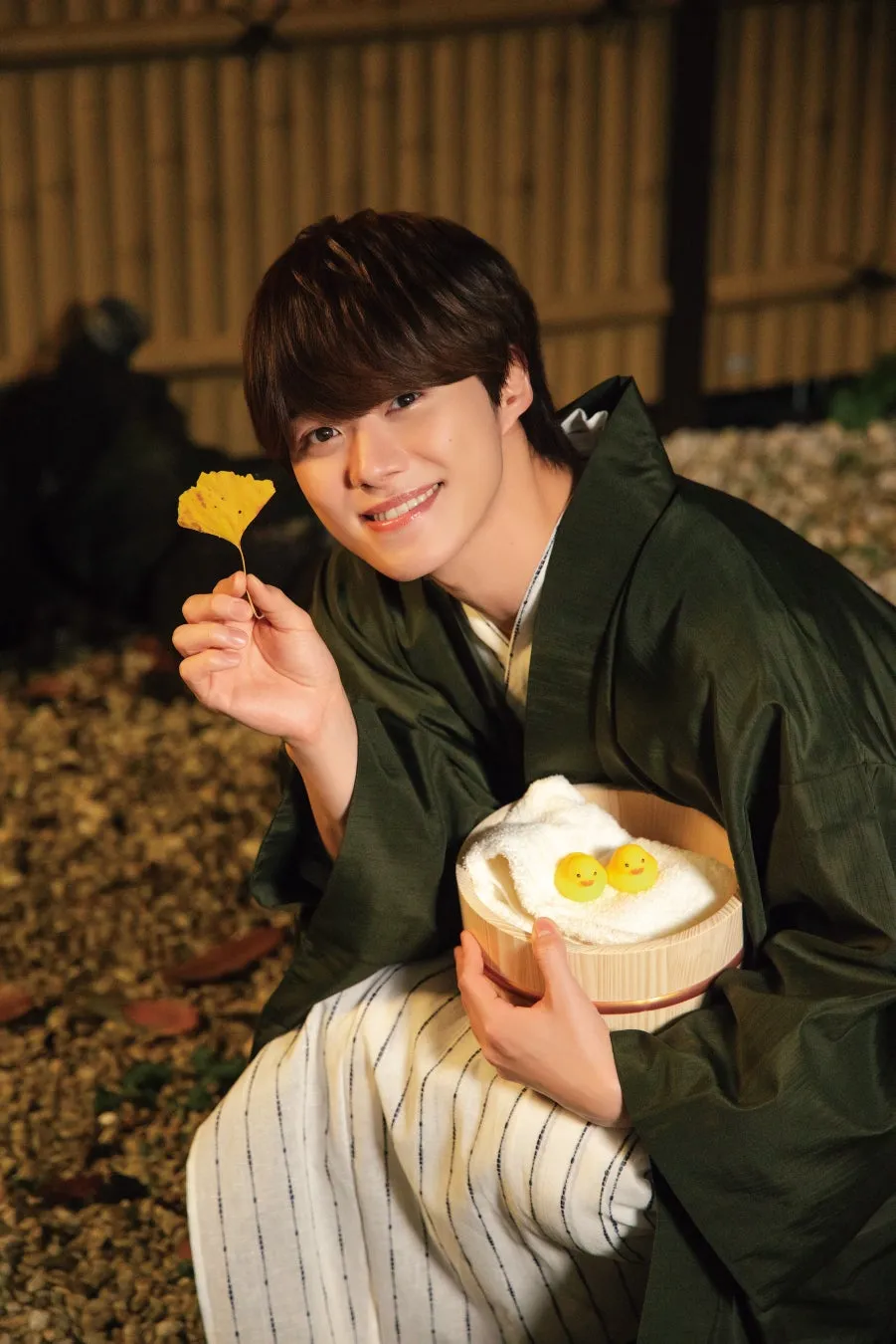 男性が浴衣姿で笑顔を見せている写真。彼は黄色い葉を持ち、手にはタオルとアヒルのおもちゃが入った桶を持っている。