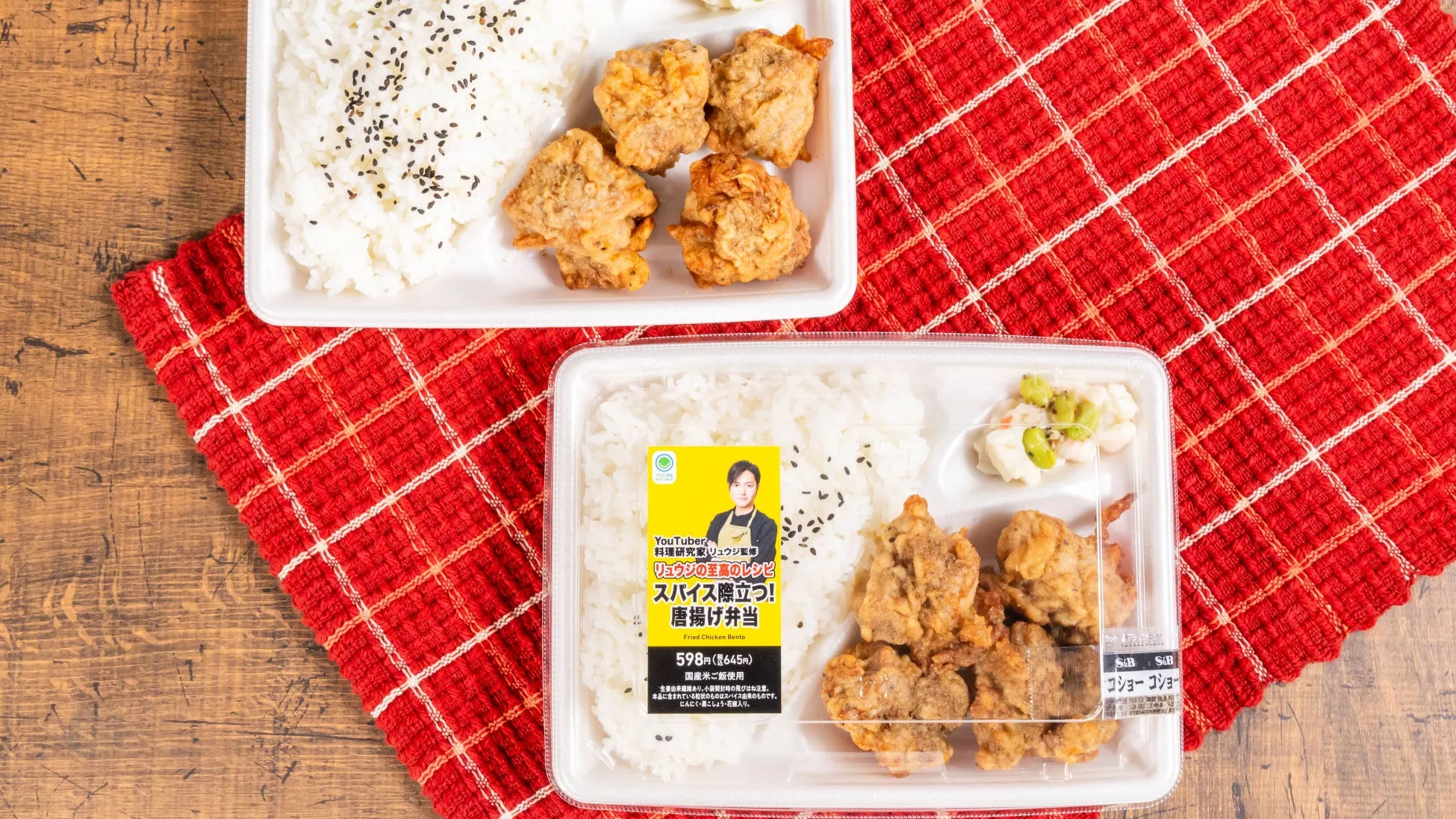 唐揚げ弁当が2つ並んだ写真。弁当にはご飯と唐揚げ、副菜が入っており、赤いチェック柄の布の上に置かれている。弁当のパッケージには「YouTuber 料理研究家 リュウジ監修」の文字が見える。