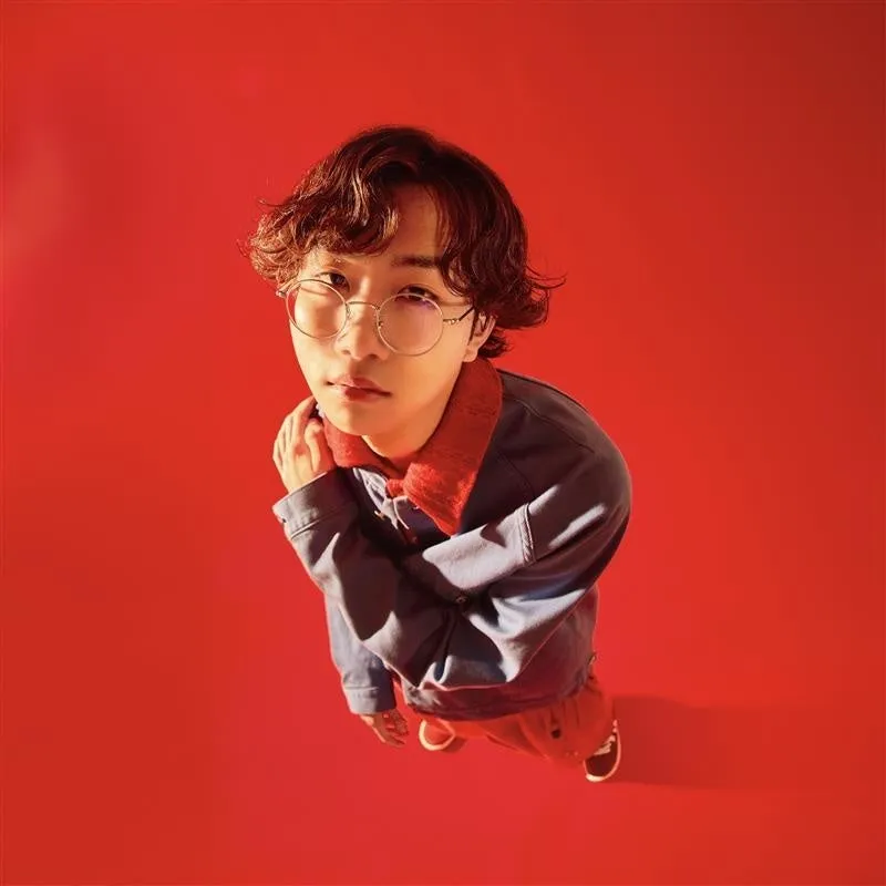 眼鏡をかけた男性が、赤い背景を前にポーズをとっている写真。彼はデニムジャケットと赤い襟のシャツを着ています。